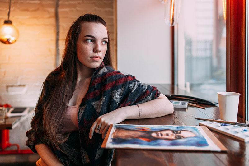 Beautiful young woman sitting by painting on table at cafe