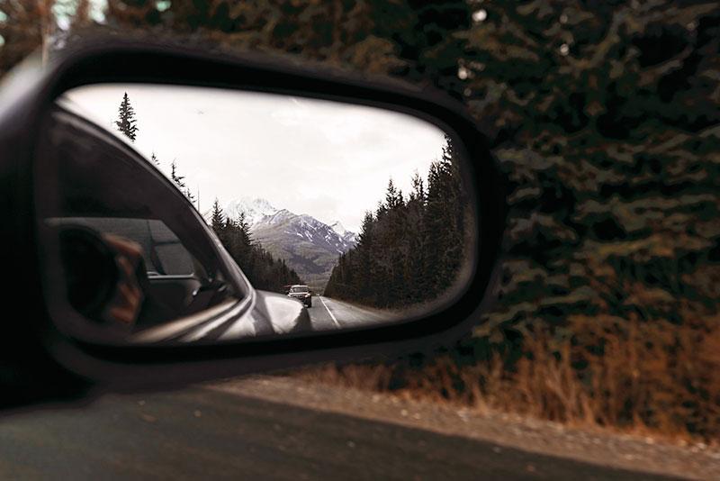 Reflection in a car mirror of mountains, caron the road and pine forest. Grey winter clouds. Cold north weather.