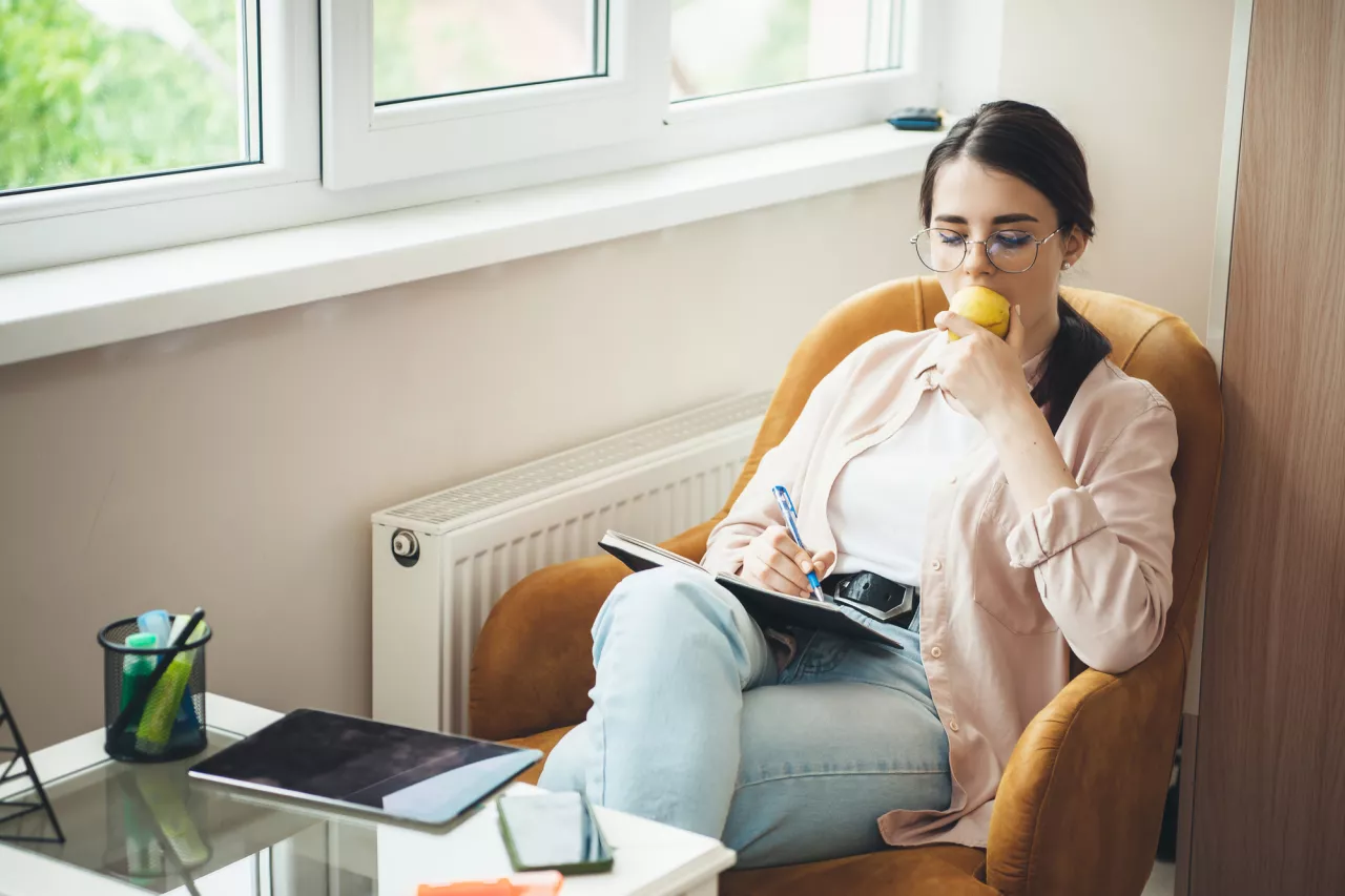 Cute caucasian lady with healthy habits is eating an apple while writing something in the book and working with a tablet