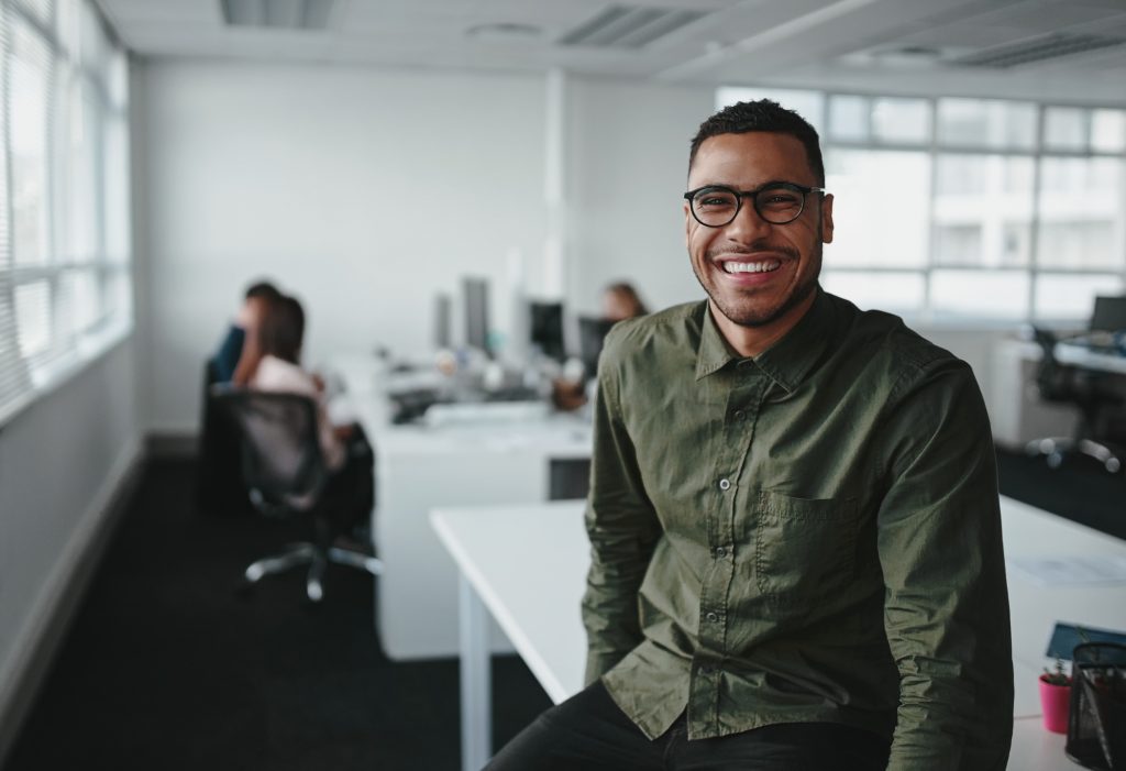 Retrato de um jovem empresário profissional sorridente bem sucedido sentado sobre a mesa na frente de um colega que trabalha em segundo plano