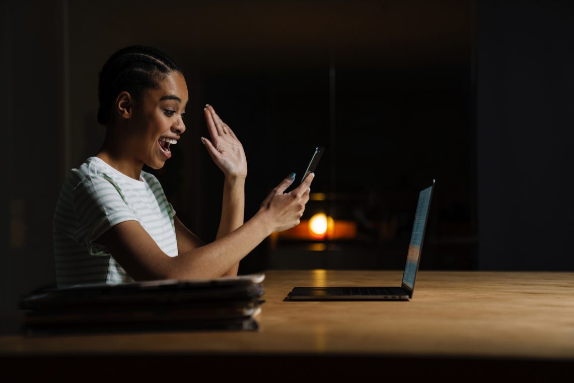 Black laughing woman waving hand and using mobile phone while in office