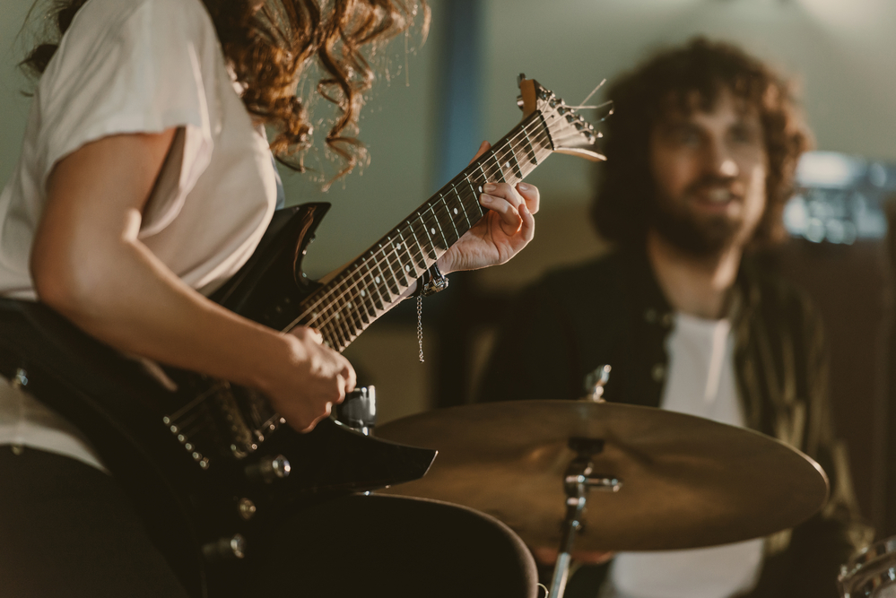 Cropped shot of female guitarist performing song with blurred drummer on background