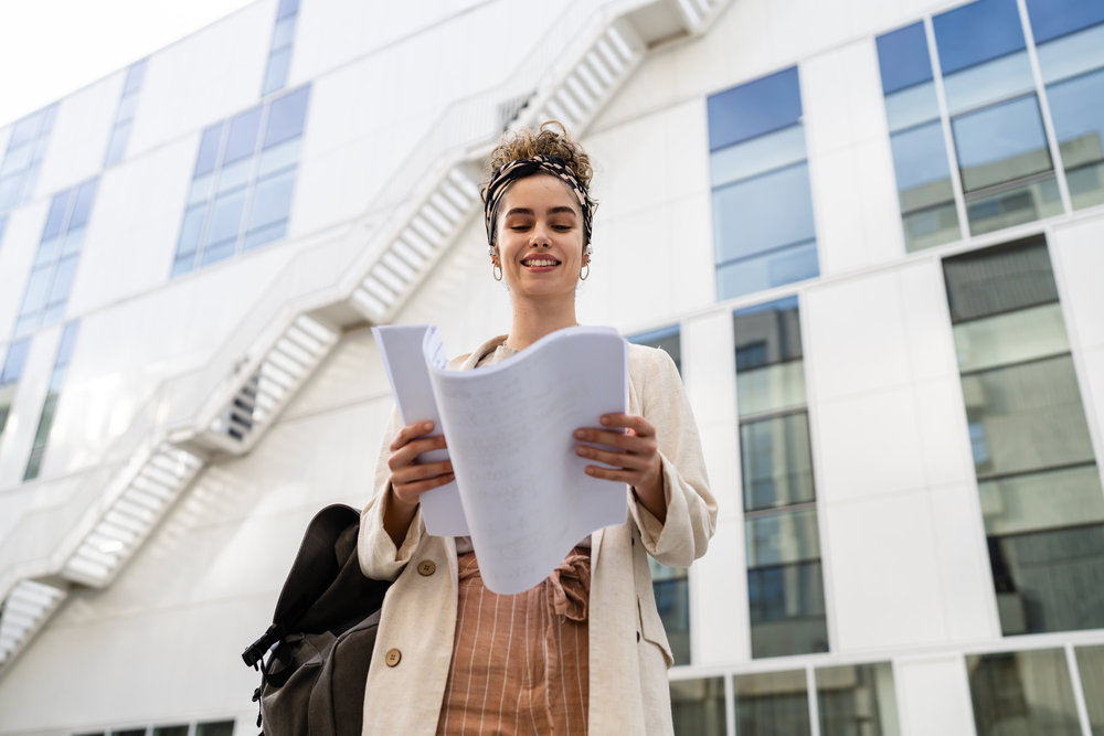 One young woman female businessman or student checking documents in front of modern building at university or company in bright day real people copy space standing alone hold paper happy smile