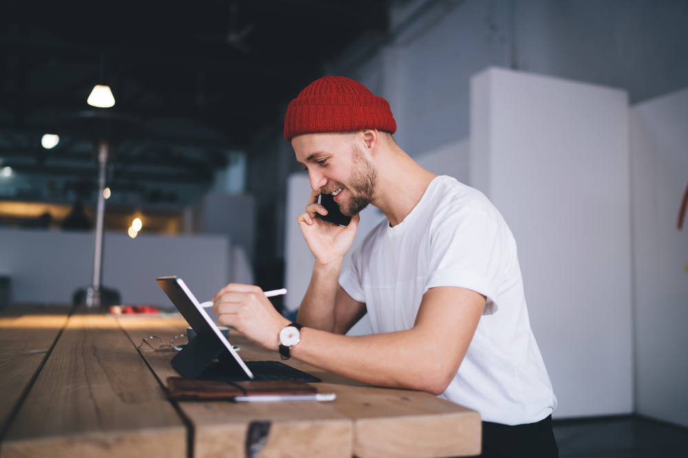 Photo side view of happy hipster adult worker in red beany sitting talking over smartphone using tablet with touchscreen pen at modern office