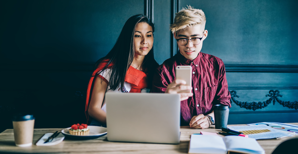 Photo Cheerful chinese friends sitting at desktop with laptop computer and tasty cake in cafe and making photo on modern smartphone device.Millennial japanese young man and woman taking pictures on cellular