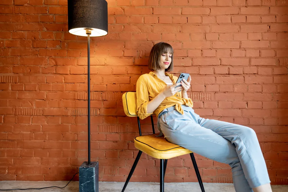 Young stylish woman using smart phone while sitting on chair near floor lamp on brick wall background