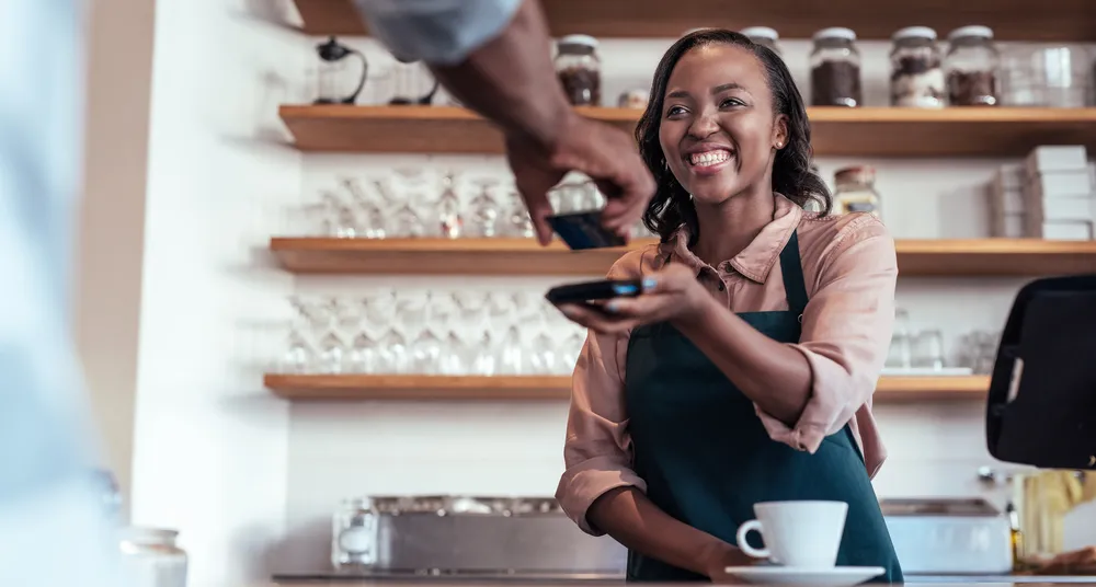 Smiling barista using nfs technology to help a customer pay for a purchase with their bank card in a cafe