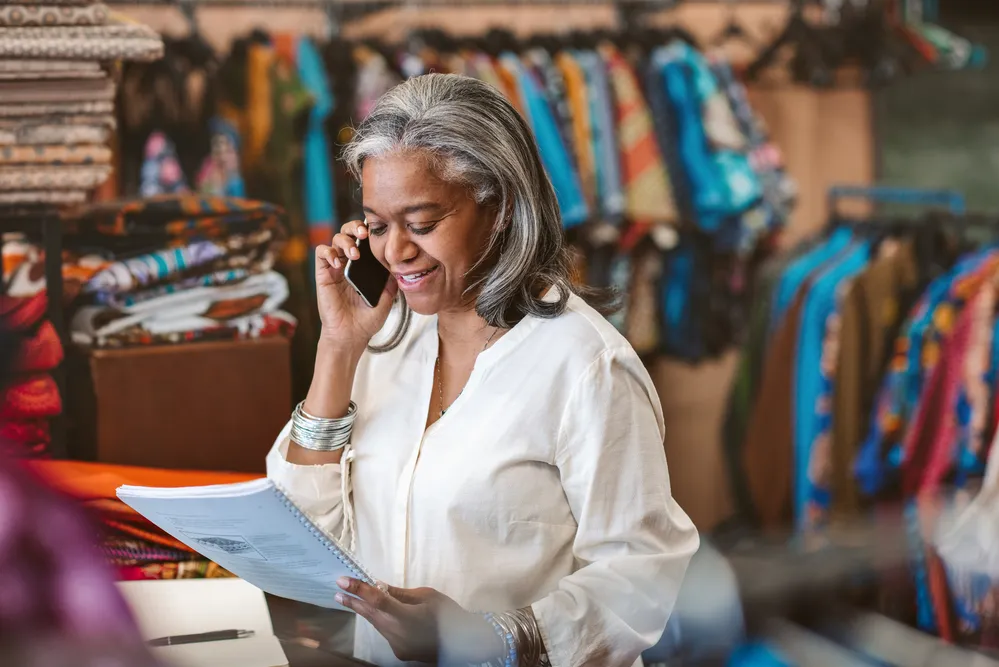 Smiling mature fabric shop owner reading paperwork and talking on a cellphone while standing behind a counter surrounded by colorful textiles