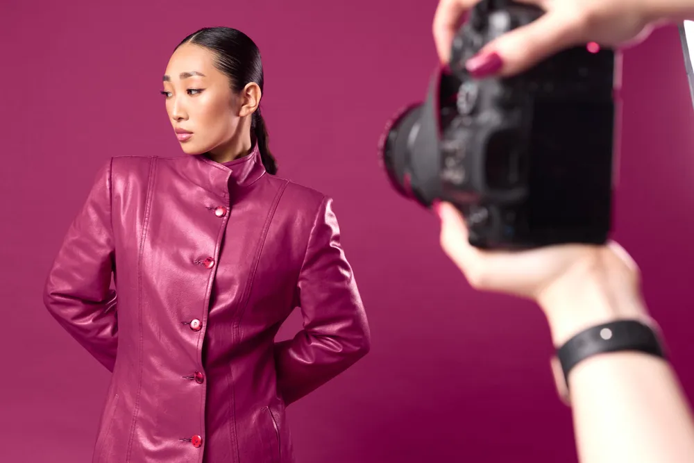 Fashion asian brunette taking photo of another woman in pink dress against purple background. woman in purple leather jacket holding camera.