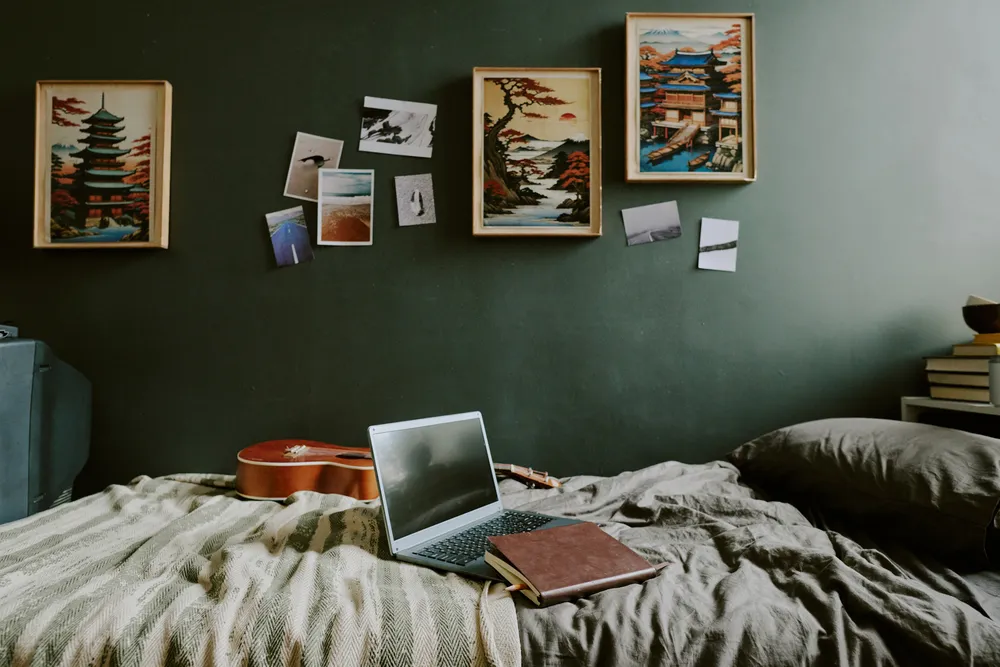 Black laptop, leather notebook and guitar on single bed made with gray linen, dark green colored wall decorated with Japan pictures and trip photos