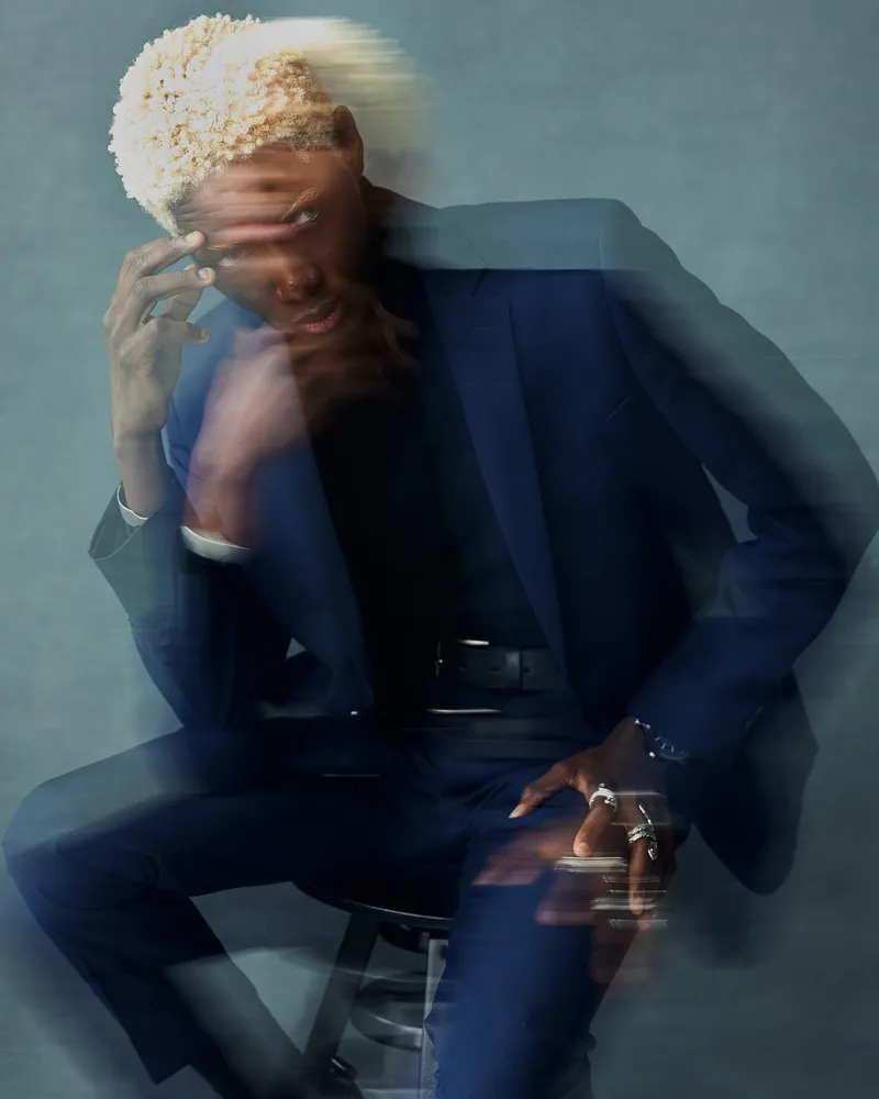 Photo of a guy sitting on a chair in a navy blue suitHe has white hair and blue eyesHe is wearing jewelryHe holds his hand to his head and looks downDouble exposure used